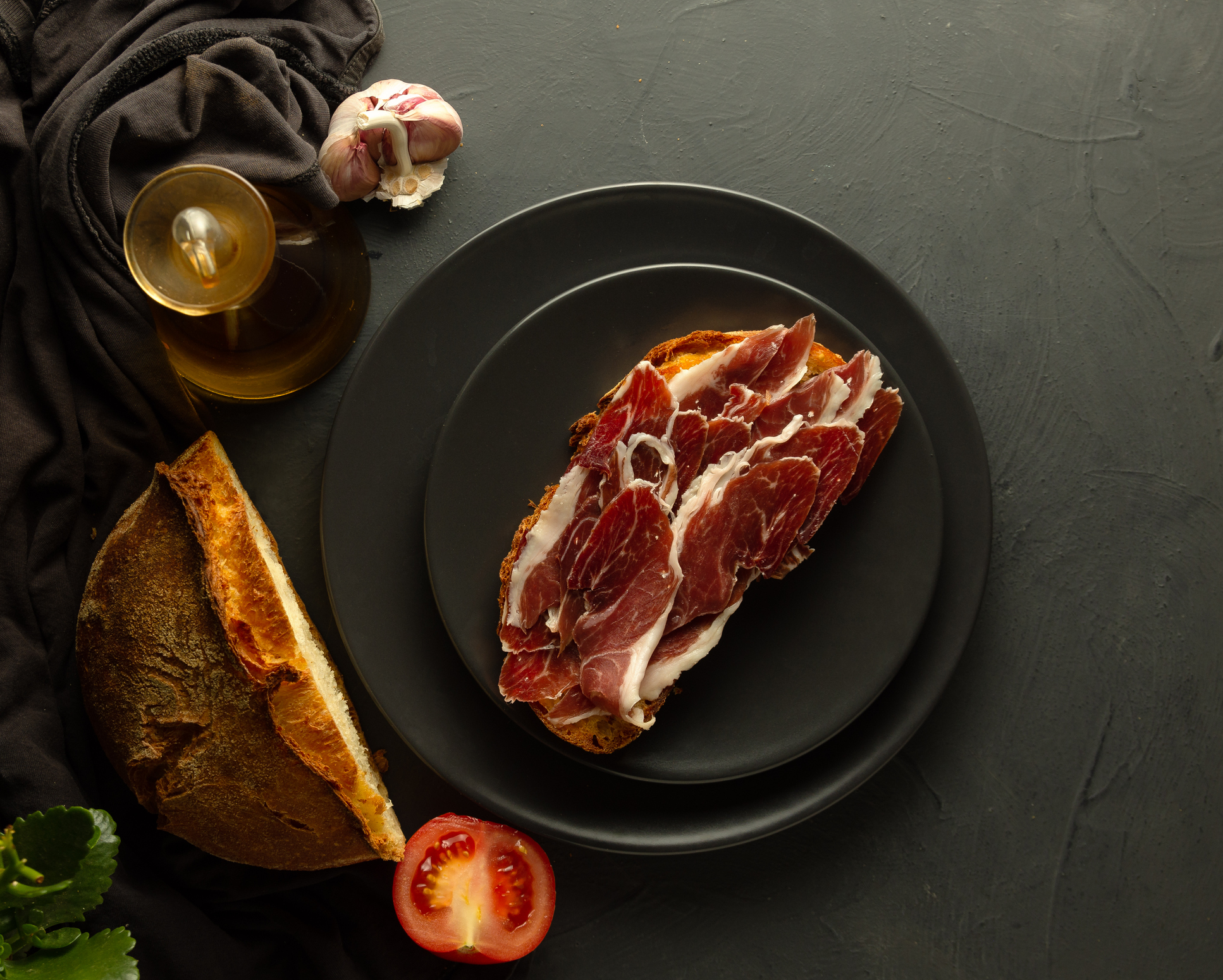 Toasted bread with Iberian ham on black plates and rustic background, in the background artisan bread, fresh tomato and olive oil